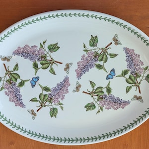 May include: An oval serving platter with a white background, featuring a lilac and butterfly design. The lilac blooms are purple, and the butterflies are blue and tan. The rim is decorated with a green leaf pattern.