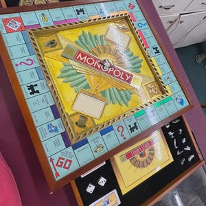Op de afbeelding: Een Monopoly-bordspelset met een houten frame, een centraal speelbord met eigendomsvakken en een uitschuifbare lade met metalen pionnen, witte dobbelstenen en kaarten. Het bord toont het Monopoly-logo en klassieke spelafbeeldingen.
