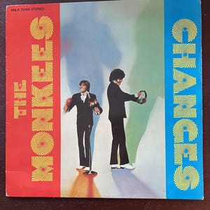 Puede incluir: Una portada de disco de vinilo roja y azul con un diseño en espiral amarillo. La portada presenta el texto "The Monkees" y "Changes" en amarillo. Dos hombres con trajes negros están de pie frente a un fondo blanco.