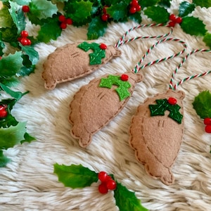 May include: Three handmade felt Christmas ornaments shaped like pastries, each decorated with green holly leaves and red berries. The ornaments are light brown and suspended by red, white, and green striped cords. The background features holly and berries.