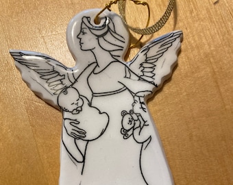 May include: White porcelain angel ornament with black line drawings of an angel holding a baby and a teddy bear. The ornament has a gold string for hanging.