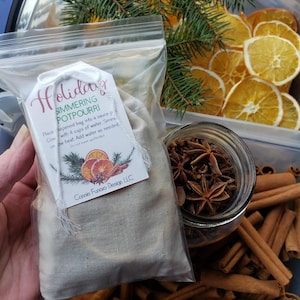 May include: A clear plastic bag containing a beige fabric bag labeled "Holiday Simmering Potpourri." The tag features illustrations of orange slices, cinnamon sticks, and pine. Beside it are dried orange slices, star anise, and cinnamon sticks, suggesting a festive theme.