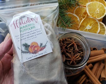 Może przedstawiać: Przezroczysta plastikowa torba zawierająca beżową torbę materiałową z napisem "Holiday Simmering Potpourri". Etykieta zawiera ilustracje plasterków pomarańczy, lasek cynamonu i sosny. Obok suszone plasterki pomarańczy, anyż gwiazdkowaty i laski cynamonu.