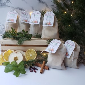 May include: Christmas simmering potpourri bags with tags, displayed on a wooden crate with dried fruit, cinnamon sticks, and greenery. The tags have floral designs and text. The bags are filled with potpourri.