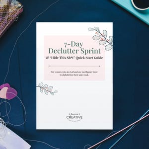 Peut inclure: Une composition à plat avec un guide blanc intitulé "7-Day Declutter Sprint & Hide This Sh*t Quick Start Guide" avec des accents floraux. D'autres articles incluent un portefeuille en peau de serpent, des lunettes de soleil et du matériel de couture sur une surface bleu foncé.