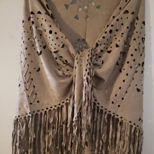 May include: A beige suede shawl with intricate cut-out designs and fringe detailing. The shawl features a knotted center and decorative cut-outs in various shapes, including circles and floral patterns. The bottom edge is adorned with long, flowing fringe, adding texture and movement to the garment.