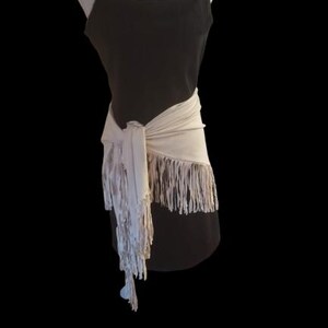 May include: A black spaghetti strap dress with a cream-colored fringed wrap tied around the waist. The wrap has long fringe that hangs down the front of the dress. The dress is displayed on a mannequin against a black background.