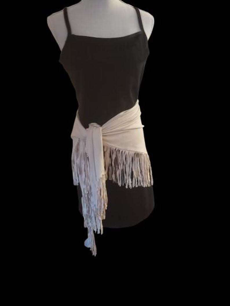May include: A black spaghetti strap dress with a cream-colored fringed wrap tied around the waist. The wrap has long fringe that hangs down the front of the dress. The dress is displayed on a mannequin against a black background.