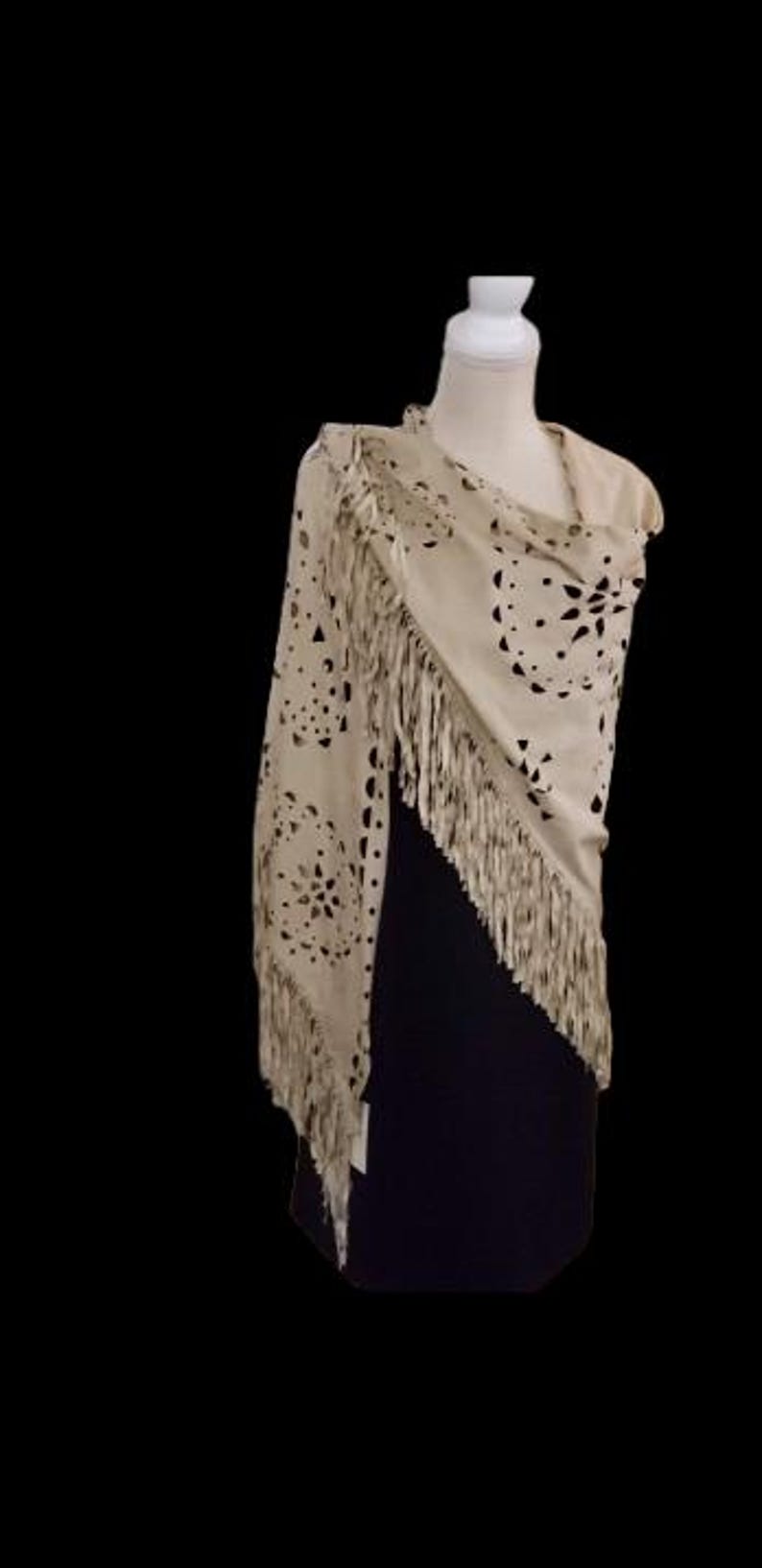 May include: A beige suede shawl with a floral cut-out design and long fringe. The shawl is draped over a mannequin, showcasing its shape and texture. The background is black, highlighting the shawl's details.