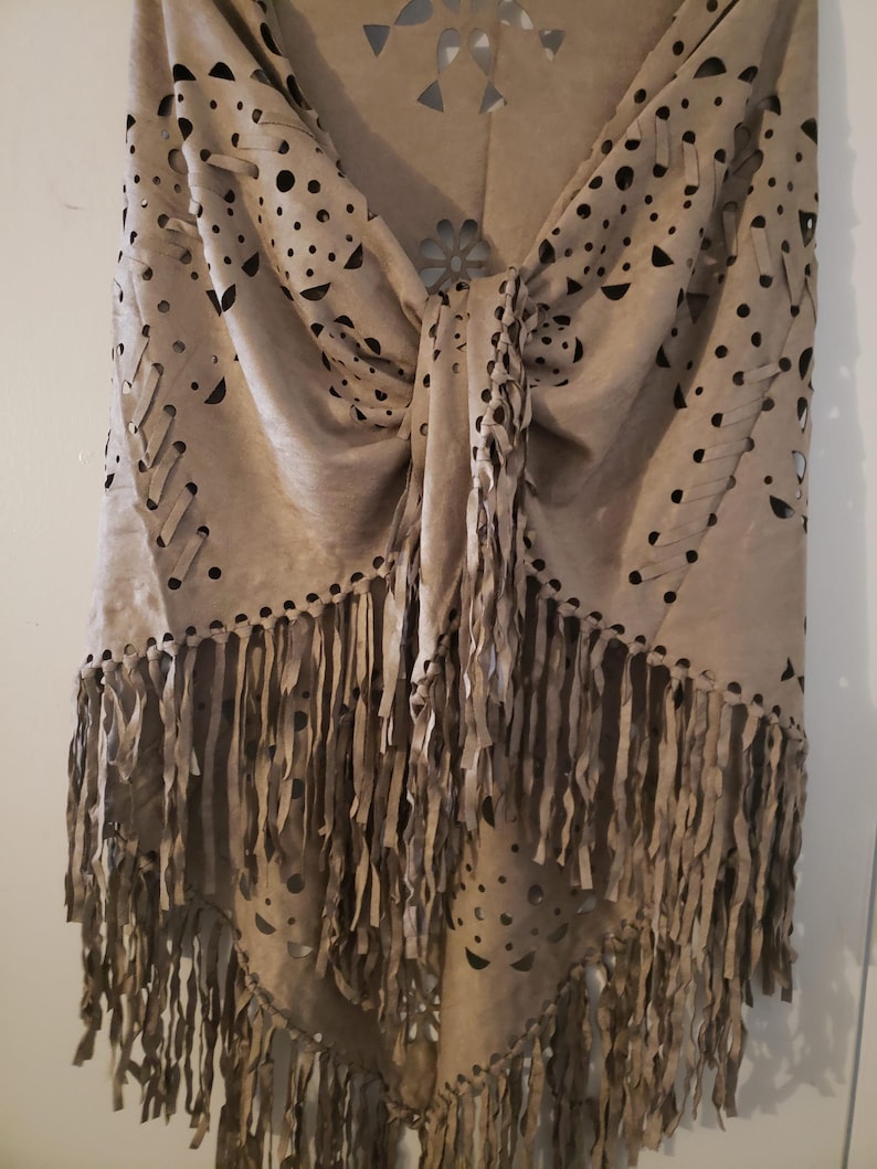 May include: A beige suede shawl with intricate cut-out designs and fringe detailing. The shawl features a knotted center and decorative cut-outs in various shapes, including circles and floral patterns. The bottom edge is adorned with long, flowing fringe, adding texture and movement to the garment.