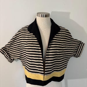 May include: A short-sleeved cropped cardigan featuring horizontal black and beige stripes. The garment has a solid black wide collar, cuffed sleeves, and a single horizontal yellow stripe near the bottom hem, finished with a solid black hemline.