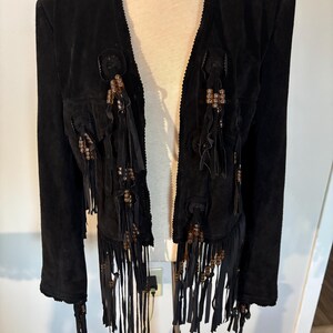 May include: Black suede jacket with fringe and beaded accents. The jacket features long sleeves and a notched collar. Decorative tassels and embellishments are sewn onto the front of the jacket. The jacket is displayed on a mannequin.