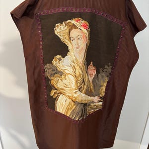 May include: Brown vest with a vertical striped pattern. The vest features a tapestry of a woman in a hat holding a container. The tapestry is framed with a burgundy border. The vest is displayed on a mannequin.