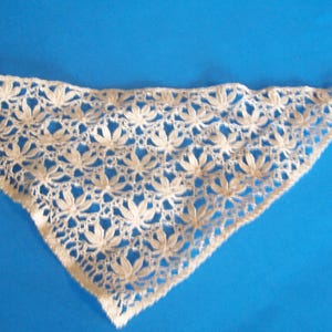 May include: Ivory-colored, triangular lace shawl with a floral pattern. The delicate lace features a repeating design of stylized flowers, creating an openwork effect. The shawl is set against a bright blue background.