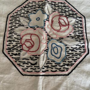 May include: A white fabric with an embroidered design of three flowers in an octagon shape. The flowers are pink, blue, and white with black outlines. The background is a black and white wavy pattern.