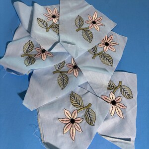 May include: A collection of light blue fabric scraps, each featuring an embroidered floral motif. The embroidery consists of a pale pink flower with a dark center and green leaves on a stem. The fabric pieces are irregularly shaped and layered.