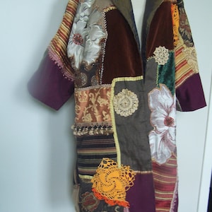 May include: A patchwork coat featuring a variety of fabrics in shades of brown, burgundy, and gold. The coat has a floral design, with a mix of textures including velvet, satin, and lace. The coat is displayed on a mannequin.