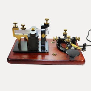 May include: A vintage telegraph key mounted on a polished dark wood base. The device features black metal components, brass adjustment screws, a silver-toned metal arm, and a black circular knob. A black electrical cord extends from the side.