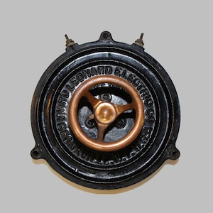 May include: A circular black metal Ward Leonard Electric Co. device from Bronxville, N.Y. featuring a central copper-colored three-spoke wheel. The housing has raised lettering around the perimeter and two small metal terminals at the top edge.