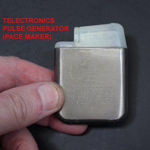 May include: A silver metal pulse generator with the text "TELETRONICS PULSE GENERATOR (PACE MAKER)" on the front. The device has a white plastic top.