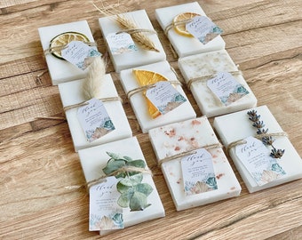 May include: Twelve white rectangular soap bars with various dried botanicals and a small thank you tag attached to each. The botanicals include dried orange slices, lavender sprigs, wheat stalks, and eucalyptus leaves. The soap bars are arranged on a wooden surface.