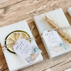 May include: White favour boxes with thank you tags tied with twine and decorated with dried fruit, wheat, and a tropical leaf design. The tags say "Thank you for celebrating this special day with us" and include the names Ainsley & Michael and the date 03 04 2020.