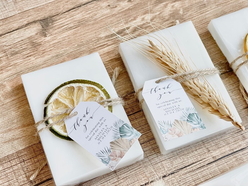 May include: White favour boxes with thank you tags tied with twine and decorated with dried fruit, wheat, and a tropical leaf design. The tags say "Thank you for celebrating this special day with us" and include the names Ainsley & Michael and the date 03 04 2020.