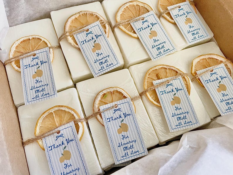 Pode incluir: Um conjunto de sabonetes retangulares brancos, cada um com uma fatia de citrino seca e uma etiqueta de papel presa com cordel. As etiquetas apresentam riscas azuis, uma ilustra&ccedil;&atilde;o de duas peras e o texto Thank You For Showering Elliott with Love.