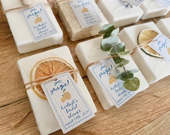 May include: Rectangular white soap bars wrapped in clear plastic, each adorned with a dried citrus slice or a sprig of eucalyptus. A rectangular paper tag with blue stripes and the text 'grazie!' and 'harleigh's bridal shower august 17th 2024' is tied on.