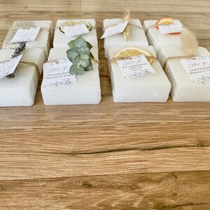 May include: Ten small white square boxes with different natural elements on top, such as dried flowers, lemons, and eucalyptus. Each box has a small thank you tag attached with text that says "Thank you for celebrating with us" or "From Rachel's shower to yours".
