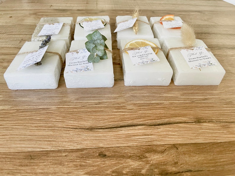 May include: Ten small white square boxes with different natural elements on top, such as dried flowers, lemons, and eucalyptus. Each box has a small thank you tag attached with text that says "Thank you for celebrating with us" or "From Rachel's shower to yours".
