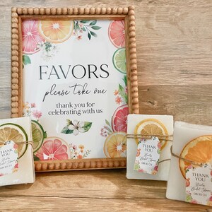 Peut inclure: Un panneau encadr&eacute; avec un motif de fruits d'agrumes indique "FAVORS please take one thank you for celebrating with us". Trois petits savons blancs sont envelopp&eacute;s de ficelle et de tranches d'agrumes s&eacute;ch&eacute;es. Chaque barre porte une petite &eacute;tiquette qui indique "THANK YOU" avec un motif floral.