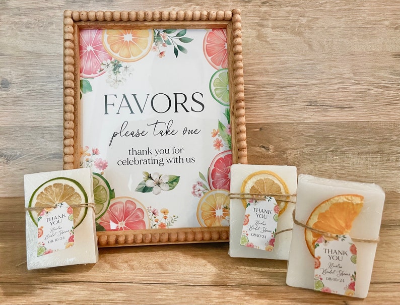 Peut inclure: Un panneau encadr&eacute; avec un motif de fruits d'agrumes indique "FAVORS please take one thank you for celebrating with us". Trois petits savons blancs sont envelopp&eacute;s de ficelle et de tranches d'agrumes s&eacute;ch&eacute;es. Chaque barre porte une petite &eacute;tiquette qui indique "THANK YOU" avec un motif floral.