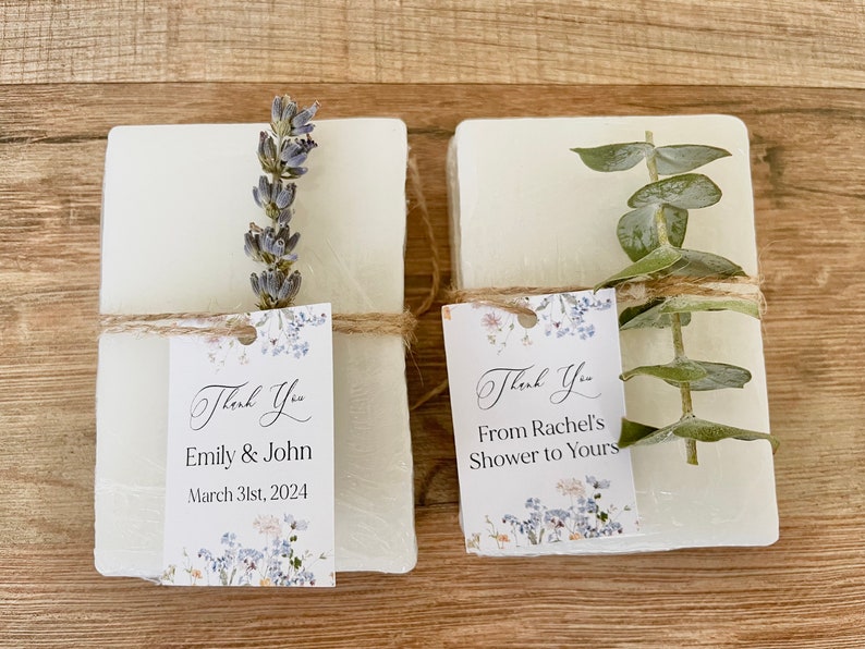 May include: Two white soap bars with floral-patterned thank you tags. One tag says "Thank You Emily & John March 31st, 2024" and the other says "Thank You From Rachel's Shower to Yours".