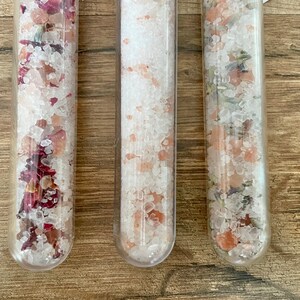 May include: Three glass test tubes filled with bath salts. The test tube on the left contains pink Himalayan salt and dried rose petals. The middle test tube contains pink Himalayan salt. The test tube on the right contains pink Himalayan salt and dried lavender.