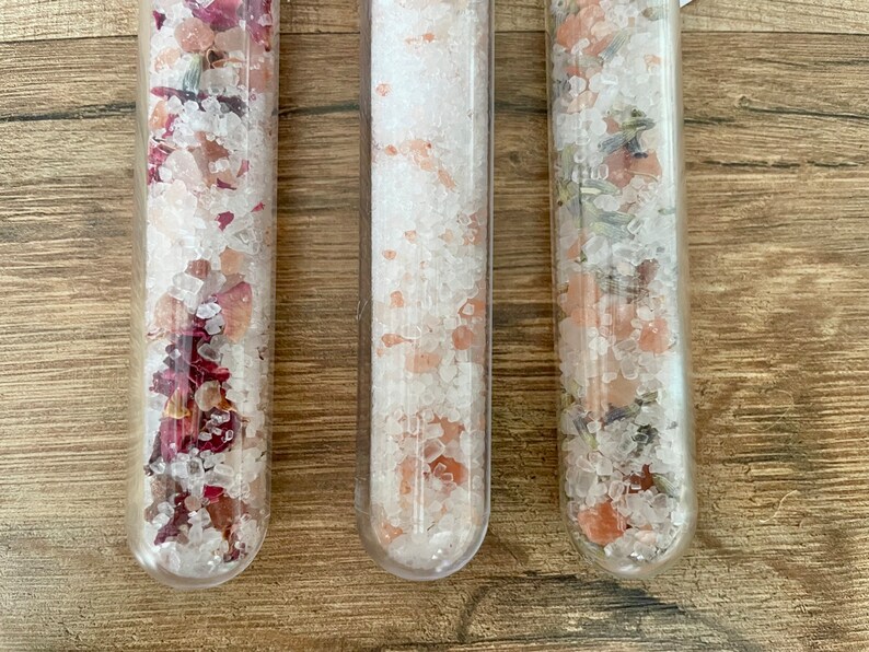 May include: Three glass test tubes filled with bath salts. The test tube on the left contains pink Himalayan salt and dried rose petals. The middle test tube contains pink Himalayan salt. The test tube on the right contains pink Himalayan salt and dried lavender.