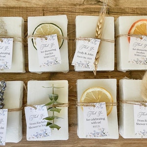May include: Eight small white soap bars wrapped in twine and decorated with dried flowers, fruit slices, and sprigs of greenery. Each bar has a small thank you tag with text that includes the names Emily & John and the date March 31, 2024.
