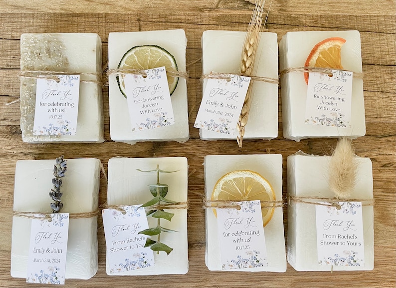 May include: Eight small white soap bars wrapped in twine and decorated with dried flowers, fruit slices, and sprigs of greenery. Each bar has a small thank you tag with text that includes the names Emily & John and the date March 31, 2024.