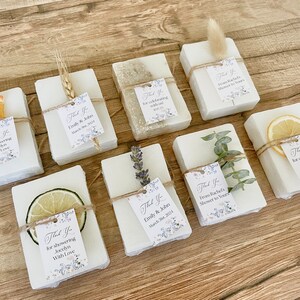 May include: A collection of small white soap bars wrapped in twine and decorated with dried flowers, fruit slices, and sprigs of greenery. Each bar has a small tag attached with the text "Thank You" and a personalized message.