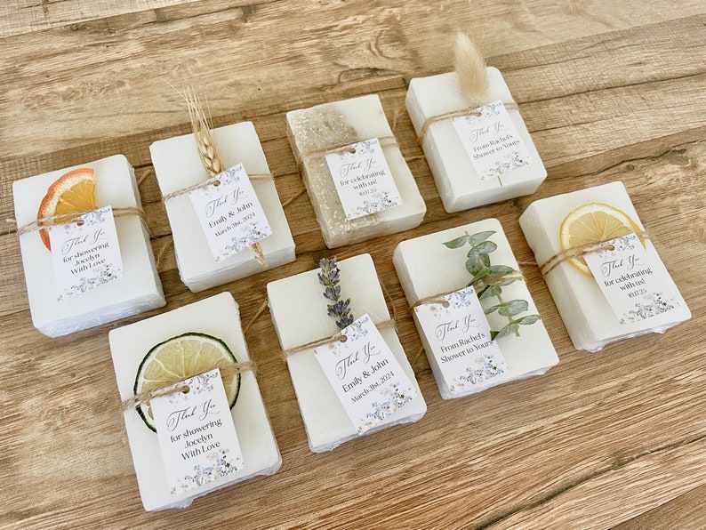 May include: A collection of small white soap bars wrapped in twine and decorated with dried flowers, fruit slices, and sprigs of greenery. Each bar has a small tag attached with the text "Thank You" and a personalized message.