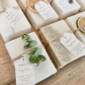 May include: Rectangular white soap bars wrapped in twine, each featuring a tag that reads AUBREY'S baptism THANK YOU FOR CELEBRATING WITH US APRIL 14TH 2024. Some bars are decorated with dried orange slices, green eucalyptus sprigs, or lavender.