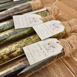 May include: Clear glass tubes filled with various dried herbs and flowers, sealed with corks and twine. Each tube has a tag that reads "THANK YOU" and "ANNA'S BRIDAL TEA 04.15.25". The tubes are arranged on a wooden surface.