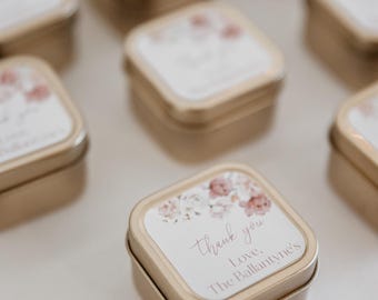 May include: Small, square, gold-colored tins with white labels featuring floral designs and the words "thank you." The tins are arranged on a white surface, suggesting a wedding or event favor.
