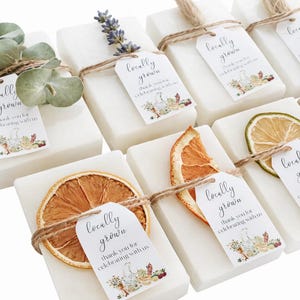 May include: A collection of rectangular white soap bars, each wrapped in twine and adorned with a tag reading locally grown thank you for celebrating with us. Some bars feature dried orange slices, lavender sprigs, or green eucalyptus leaves.