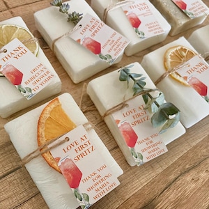May include: Rectangular white soap bars decorated with dried citrus slices, lavender sprigs, or eucalyptus leaves. Each bar features a tied tag with twine, displaying the text "LOVE AT FIRST SPRITZ" and "THANK YOU FROM REBECCA'S SHOWER TO YOURS" or "MARIA WITH LOVE."