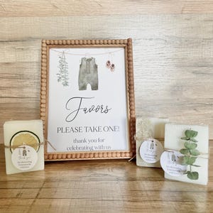 May include: A wooden beaded frame containing a sign that reads Favors PLEASE TAKE ONE! thank you for celebrating with us, featuring illustrations of a baby romper, shoes, and greenery. Three rectangular soap bars with decorative accents sit in front.