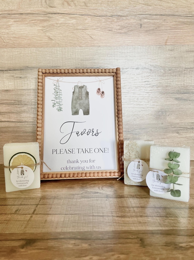 May include: A wooden beaded frame containing a sign that reads Favors PLEASE TAKE ONE! thank you for celebrating with us, featuring illustrations of a baby romper, shoes, and greenery. Three rectangular soap bars with decorative accents sit in front.