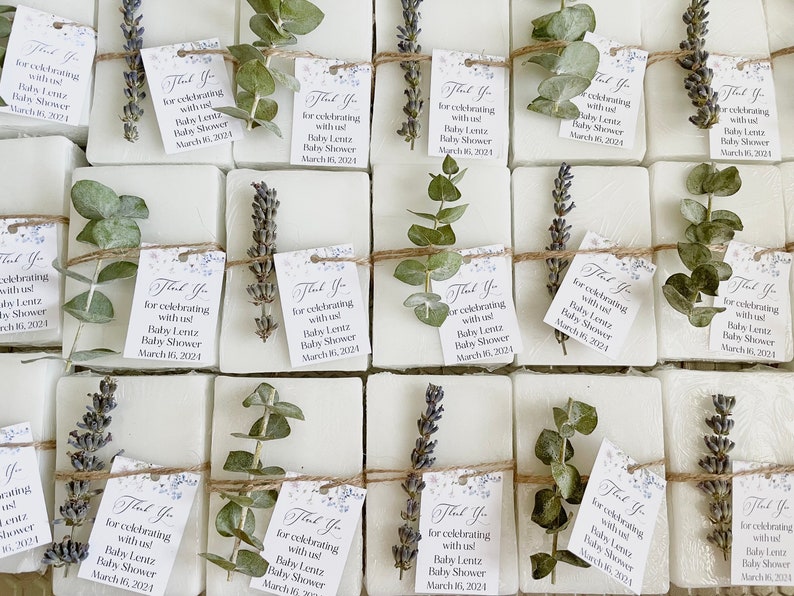 May include: A collection of small white soap bars wrapped in twine and decorated with sprigs of eucalyptus and lavender. Each bar has a small tag attached that reads "Thank You for celebrating with us! Baby Lentz Baby Shower March 16, 2024".