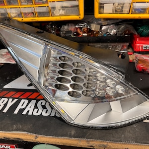 May include: A silver and black car taillight with a honeycomb pattern and a silver trim. The taillight is on a black surface with the text "HOBBY PASSION" visible.