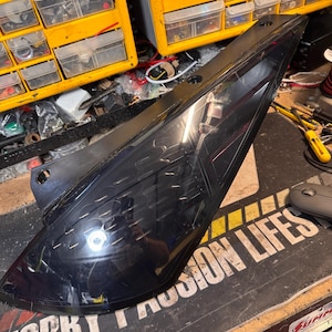 May include: Black automotive taillight with a smoked lens. The taillight is on a workbench with a yellow and black caution tape mat and a mouse.